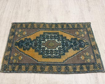 Puede incluir: Una alfombra rectangular que presenta un medallón central en forma de diamante en amarillo mostaza y azul oscuro. La alfombra tiene un fondo marrón con un patrón de borde floral repetitivo en azul y amarillo, mostrando un diseño geométrico.