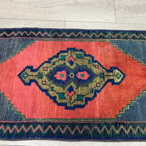 K&ouml;nnte beinhalten: Rechteckiger Teppich mit einem rautenf&ouml;rmigen Mitteldesign in Koralle und einem marineblauen Rand. Die Mitte weist ein geometrisches Muster in Gold, T&uuml;rkis und Marineblau auf. Der Teppich hat einen Fransenrand und einen Zickzack-Rand.