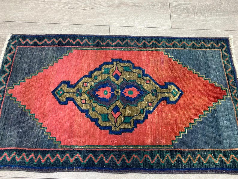K&ouml;nnte beinhalten: Rechteckiger Teppich mit einem rautenf&ouml;rmigen Mitteldesign in Koralle und einem marineblauen Rand. Die Mitte weist ein geometrisches Muster in Gold, T&uuml;rkis und Marineblau auf. Der Teppich hat einen Fransenrand und einen Zickzack-Rand.