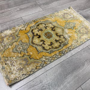 Puede incluir: Alfombra rectangular de estilo vintage con un dise&ntilde;o de medall&oacute;n central. La alfombra presenta un aspecto envejecido con una paleta de colores amarillo, verde y marr&oacute;n. Se encuentra sobre un suelo de madera gris.
