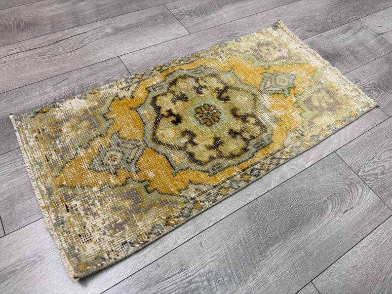Puede incluir: Alfombra rectangular de estilo vintage con un dise&ntilde;o de medall&oacute;n central. La alfombra presenta un aspecto envejecido con una paleta de colores amarillo, verde y marr&oacute;n. Se encuentra sobre un suelo de madera gris.