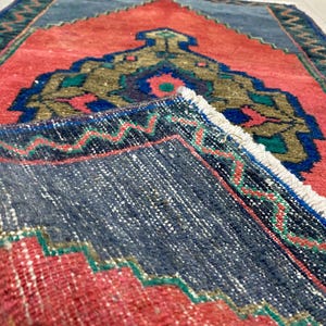 K&ouml;nnte beinhalten: Ein gefalteter, rechteckiger Teppich mit einem lebendigen Design. Der Teppich hat ein korallenfarbenes Feld mit einem zentralen, verzierten Medaillon in Gold-, Gr&uuml;n- und Blaut&ouml;nen. Ein dunkelblauer Rand mit Zickzackmuster umrahmt den Teppich, und die Kanten haben einen Fransen.