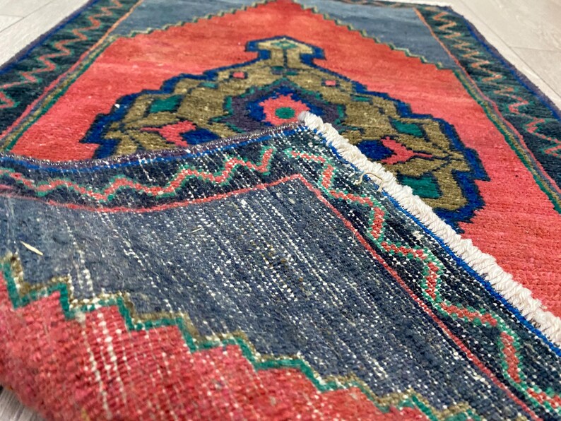K&ouml;nnte beinhalten: Ein gefalteter, rechteckiger Teppich mit einem lebendigen Design. Der Teppich hat ein korallenfarbenes Feld mit einem zentralen, verzierten Medaillon in Gold-, Gr&uuml;n- und Blaut&ouml;nen. Ein dunkelblauer Rand mit Zickzackmuster umrahmt den Teppich, und die Kanten haben einen Fransen.