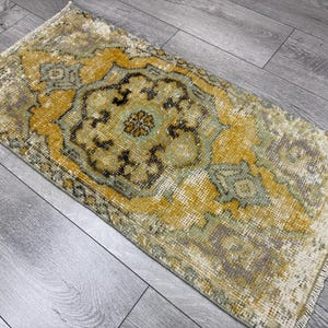 Puede incluir: Alfombra rectangular de estilo vintage con un dise&ntilde;o central de medall&oacute;n. La alfombra presenta un aspecto envejecido con una paleta de colores dorados, verde oliva y marr&oacute;n. Los bordes tienen flecos, lo que aumenta su est&eacute;tica envejecida.