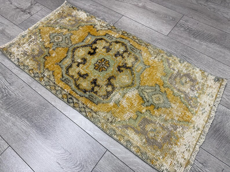 Puede incluir: Alfombra rectangular de estilo vintage con un dise&ntilde;o central de medall&oacute;n. La alfombra presenta un aspecto envejecido con una paleta de colores dorados, verde oliva y marr&oacute;n. Los bordes tienen flecos, lo que aumenta su est&eacute;tica envejecida.