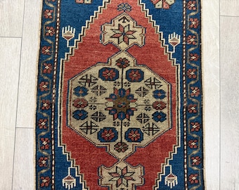 Puede incluir: Una alfombra tejida rectangular que presenta un hexágono central beige con motivos florales azules y rojos, rodeado por una gran forma de diamante roja sobre un fondo azul intenso. El borde muestra un patrón geométrico repetitivo en azul y rojo.
