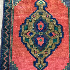 K&ouml;nnte beinhalten: Ein rechteckiger Teppich mit einem zentralen Medaillon-Design. Der Teppich hat ein korallenfarbenes Feld mit einem zentralen, verzierten Muster in Gold, Blau und Gr&uuml;n. Der Rand ist dunkelblau mit einem Zickzackmuster. Der Teppich ist wahrscheinlich aus Wolle.