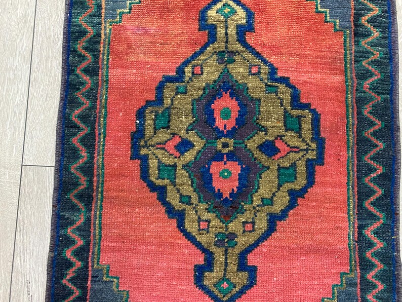 K&ouml;nnte beinhalten: Ein rechteckiger Teppich mit einem zentralen Medaillon-Design. Der Teppich hat ein korallenfarbenes Feld mit einem zentralen, verzierten Muster in Gold, Blau und Gr&uuml;n. Der Rand ist dunkelblau mit einem Zickzackmuster. Der Teppich ist wahrscheinlich aus Wolle.