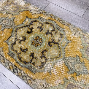Puede incluir: Una alfombra rectangular de aspecto envejecido, con un dise&ntilde;o central de medall&oacute;n. La paleta de colores de la alfombra incluye amarillo mostaza, verde salvia y gris apagado, con patrones detallados y un estilo vintage. Adecuada para la decoraci&oacute;n del hogar.