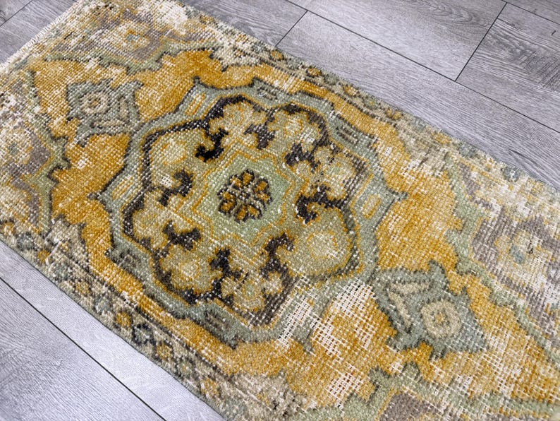 Puede incluir: Una alfombra rectangular de aspecto envejecido, con un dise&ntilde;o central de medall&oacute;n. La paleta de colores de la alfombra incluye amarillo mostaza, verde salvia y gris apagado, con patrones detallados y un estilo vintage. Adecuada para la decoraci&oacute;n del hogar.