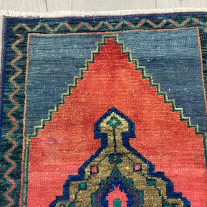 K&ouml;nnte beinhalten: Rechteckiger Teppich mit einem zentralen Design in Orange- und Blaut&ouml;nen. Der Teppich hat ein geometrisches Muster mit einem zentralen Medaillon und einem Rand mit Zickzack-Design. Der Teppich ist wahrscheinlich aus gewebtem Stoff.