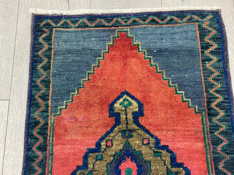 K&ouml;nnte beinhalten: Rechteckiger Teppich mit einem zentralen Design in Orange- und Blaut&ouml;nen. Der Teppich hat ein geometrisches Muster mit einem zentralen Medaillon und einem Rand mit Zickzack-Design. Der Teppich ist wahrscheinlich aus gewebtem Stoff.
