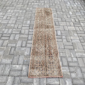 Könnte beinhalten: Ein langer, rechteckiger Teppich mit einem braunen und beigefarbenen geometrischen Muster. Der Teppich hat einen Fransenrand und liegt auf einer grauen Steinplatte.