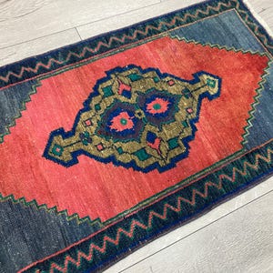 K&ouml;nnte beinhalten: Ein rechteckiger Teppich mit einem korallenfarbenen Mittelfeld und einem detaillierten, verzierten Design in Gold, Blau und T&uuml;rkis. Der Teppich hat einen dunkelblauen Rand mit einem Zickzackmuster. Die Abmessungen des Teppichs sind nicht sichtbar.