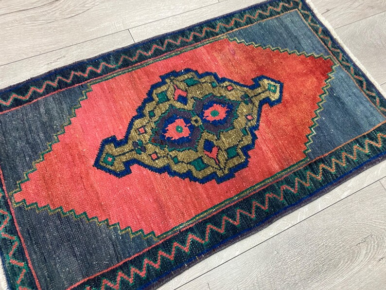 K&ouml;nnte beinhalten: Ein rechteckiger Teppich mit einem korallenfarbenen Mittelfeld und einem detaillierten, verzierten Design in Gold, Blau und T&uuml;rkis. Der Teppich hat einen dunkelblauen Rand mit einem Zickzackmuster. Die Abmessungen des Teppichs sind nicht sichtbar.