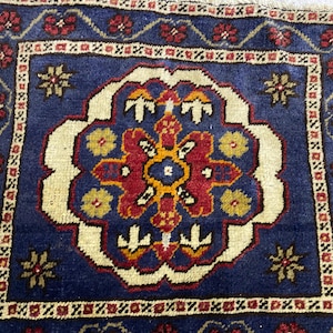 May include: A square rug featuring a deep blue field with a central cream-colored medallion containing red, orange, and yellow floral motifs. The border displays a repeating pattern of red flowers and geometric shapes on a cream background.