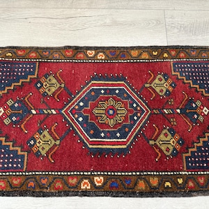May include: A rectangular woven rug featuring a deep red central field with a large navy blue hexagonal medallion containing a floral motif. The rug is decorated with four smaller floral elements in the corners and a brown border with geometric patterns.