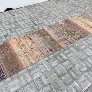 Op de afbeelding: Een vintage Turkse loper met een vervaagd, veelkleurig patroon. De loper is rechthoekig en gemaakt van wol. Hij heeft een verscheidenheid aan kleuren, waaronder rood, oranje, geel, blauw en groen. De loper is in goede staat en heeft een versleten, verweerde uitstraling.