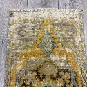 Puede incluir: Una alfombra rectangular desgastada con una apariencia descolorida y vintage. La alfombra presenta un dise&ntilde;o central en tonos dorados, verdes y grises, con patrones intrincados y un borde con flecos. La alfombra est&aacute; colocada sobre una superficie de madera gris claro.