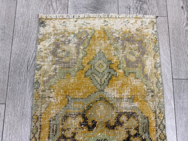 Puede incluir: Una alfombra rectangular desgastada con una apariencia descolorida y vintage. La alfombra presenta un dise&ntilde;o central en tonos dorados, verdes y grises, con patrones intrincados y un borde con flecos. La alfombra est&aacute; colocada sobre una superficie de madera gris claro.