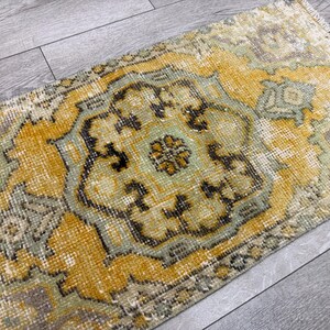 Puede incluir: Una alfombra rectangular y desgastada con un dise&ntilde;o de medall&oacute;n central. La alfombra presenta una paleta de colores amarillo, verde salvia y marr&oacute;n, con patrones intrincados y una est&eacute;tica vintage. La alfombra est&aacute; colocada sobre un suelo de madera gris.