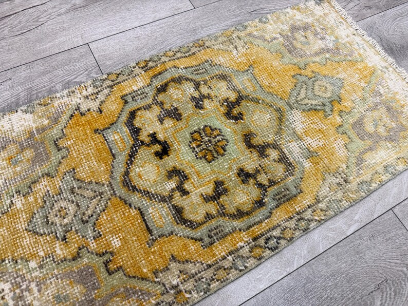 Puede incluir: Una alfombra rectangular y desgastada con un dise&ntilde;o de medall&oacute;n central. La alfombra presenta una paleta de colores amarillo, verde salvia y marr&oacute;n, con patrones intrincados y una est&eacute;tica vintage. La alfombra est&aacute; colocada sobre un suelo de madera gris.