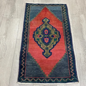 K&ouml;nnte beinhalten: Ein rechteckiger, handgewebter Teppich mit einem zentralen Rautenmuster in Koralle und Gold, umrahmt von marineblauen geometrischen Formen und einem marineblauen Rand. Die Ma&szlig;e betragen ca. 61 x 91 cm.
