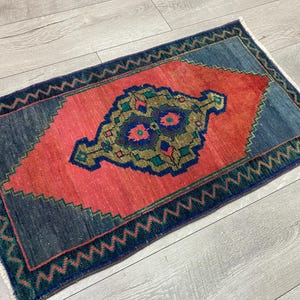 K&ouml;nnte beinhalten: Ein rechteckiger, handgewebter Teppich mit einem zentralen Medaillon-Design. Der Teppich hat eine lebendige Farbpalette, darunter ein korallenrotes Zentrum, umgeben von marineblauen und t&uuml;rkisgr&uuml;nen R&auml;ndern. Die Kanten haben ein Zickzackmuster.