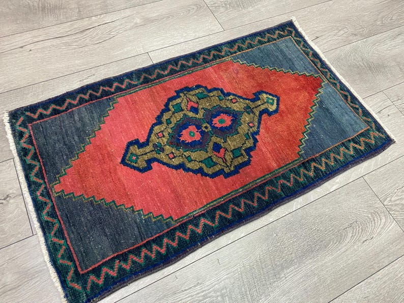 K&ouml;nnte beinhalten: Ein rechteckiger, handgewebter Teppich mit einem zentralen Medaillon-Design. Der Teppich hat eine lebendige Farbpalette, darunter ein korallenrotes Zentrum, umgeben von marineblauen und t&uuml;rkisgr&uuml;nen R&auml;ndern. Die Kanten haben ein Zickzackmuster.