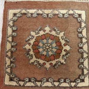 May include: A rectangular woven textile wall hanging featuring a terracotta brown base colour. The centre displays a cream-coloured floral medallion with blue and orange accents, surrounded by a cream border with a repeating tufted pattern.