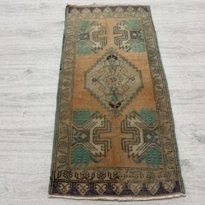 May include: A rectangular rug featuring a central diamond medallion in muted tones of tan, teal, and dark brown. The design includes geometric motifs and stylized floral patterns within a decorative border, finished with short fringe at both ends.