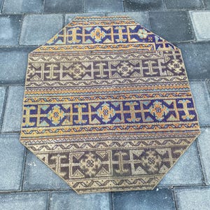 May include: An octagonal rug with a detailed, geometric pattern. The rug features a central field of brown with repeating shapes, bordered by bands of blue, orange, and yellow. The rug is placed on a gray brick surface.