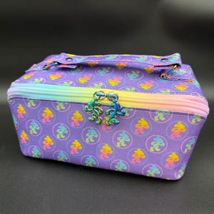 May include: A rectangular purple cosmetic bag featuring a repeating pattern of small, colorful dancing bear silhouettes inside circles. The bag has a rainbow-colored zipper closure and two iridescent metal zipper pulls shaped like dancing bears.