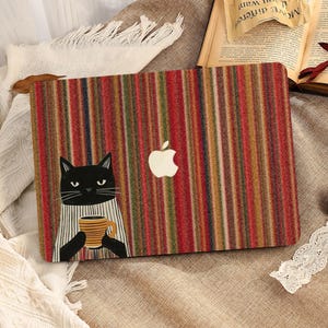 Könnte beinhalten: Laptop-Hülle mit vertikalem Streifenmuster in Rot, Braun und Beige. Auf der linken Seite befindet sich eine Illustration einer schwarzen Katze, die einen Becher hält. Das Apple-Logo ist sichtbar. Die Hülle ist für einen Laptop.