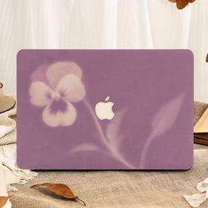 May include: A matte purple laptop hard shell case featuring a soft-focus, light-colored pansy flower graphic on the left side. The center of the case displays a cutout revealing the silver Apple logo, with a smooth, solid-colored finish.