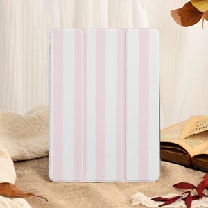 Puede incluir: Una funda para tableta con diseño de rayas blancas y rosas. La funda tiene una cubierta plegable. La funda está sobre una superficie marrón, con un libro abierto y hojas decorativas en el fondo.