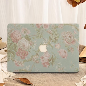 May include: A laptop case with a floral pattern. The case has a pale green base with pink and white flowers. The Apple logo is in the centre. The case is on a brown surface.