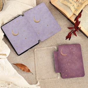 May include: Two e-reader cases in shades of purple, one light and one dark, both featuring gold crescent moon and star patterns. The light purple case is open, and the dark purple case is closed. A clear plastic protective shell is also included.