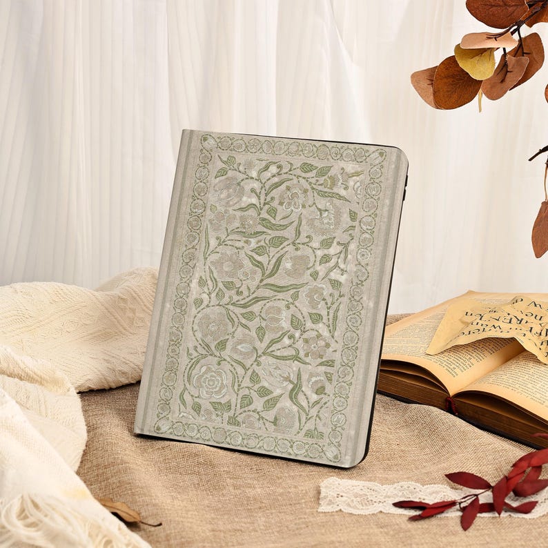 May include: A light gray book cover with a floral design in shades of green and white. The cover features a rectangular pattern of flowers and leaves, with a decorative border. The book is open to reveal pages.