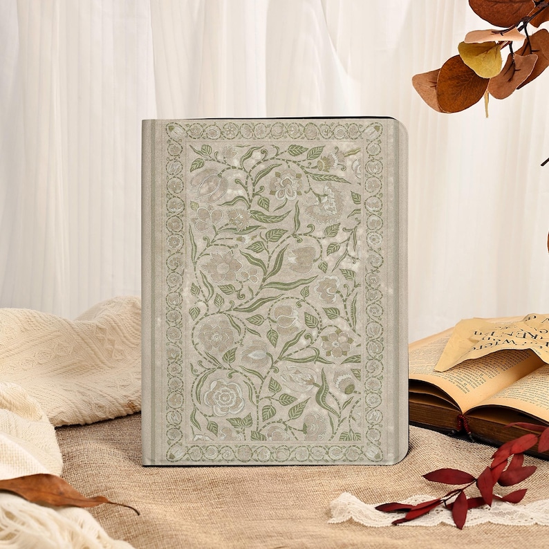 May include: A hardback journal with a floral design. The cover features a light green and white floral pattern with a decorative border. The journal is rectangular and appears to be closed. The background includes an open book and decorative elements.