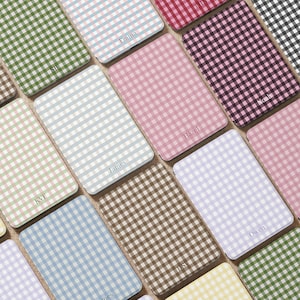 Puede incluir: Colección de posavasos rectangulares con estampado de cuadros vichy en varios colores, como rosa, verde, azul y marrón. Cada posavasos tiene un nombre o iniciales impresos en color claro.