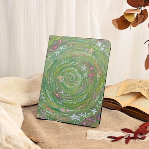 Może przedstawiać: Zielone etui na tablet z kwiatowym wzorem. Etui posiada spiralny wz&oacute;r z białymi, r&oacute;żowymi i ż&oacute;łtymi kwiatami. Tło to mieszanka jasnych i ciemnych odcieni zieleni. Etui leży na brązowej powierzchni.