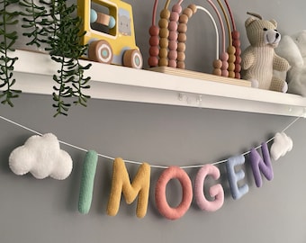 May include: A decorative felt name banner spelling IMOGEN in multi-coloured letters, including sage green, mustard yellow, coral, light pink, light blue, and purple. The banner features a white cloud at each end, all strung together on a thin white cord.