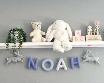 Könnte beinhalten: Ein weißes Regal trägt eine grüne Pflanze, einen beigen Teddybären, einen weißen Plüschhasen, rosa Buchstabenwürfel und ein Holzschild mit der Aufschrift bee happy. Darunter hängt eine Girlande mit zwei grauen Filzhasen und den Buchstaben NOAH.