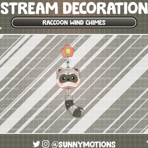 Peut inclure: Un carillon à vent en forme de raton laveur avec un visage de raton laveur gris et blanc, une fleur rose et une queue gris et blanc. Le carillon est suspendu à une ficelle blanche.