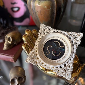 May include: A small, square, off-white ornate frame containing a dark circular display with a butterfly and a gold-colored snake. The frame features intricate scrollwork and sits atop a shiny, gold-colored hand-shaped sculpture.