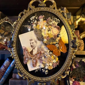 May include: An oval-shaped framed picture with a black background. The frame is gold and ornate. Inside the frame is a black and white photograph of a man wearing a suit and tie. The photograph is surrounded by dried flowers and butterflies.