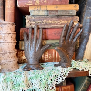 May include: Two dark brown, cast iron hand-shaped sculptures with visible finger details and mounting bases. The hands are positioned upright, facing forward, and feature a textured, weathered finish. They are displayed in front of a stack of vintage books.