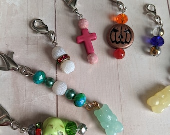 May include: A collection of colorful beaded charms with different designs, including a cross, a pumpkin, a skull, a bear, and a variety of other shapes. The charms are attached to silver clasps and are perfect for adding a touch of personality to your projects.
