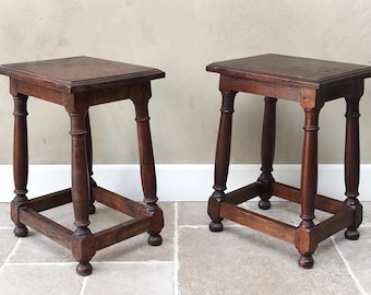 May include: Two antique wooden stools with square seats and turned legs. The stools are a dark brown color and have a simple, classic design. The legs are connected by a square base. The stools are likely vintage or antique furniture.